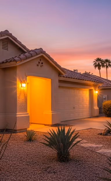 Single-story house with tiled roof and lit entrance and garage at sunset, with desert landscaping and palm trees.